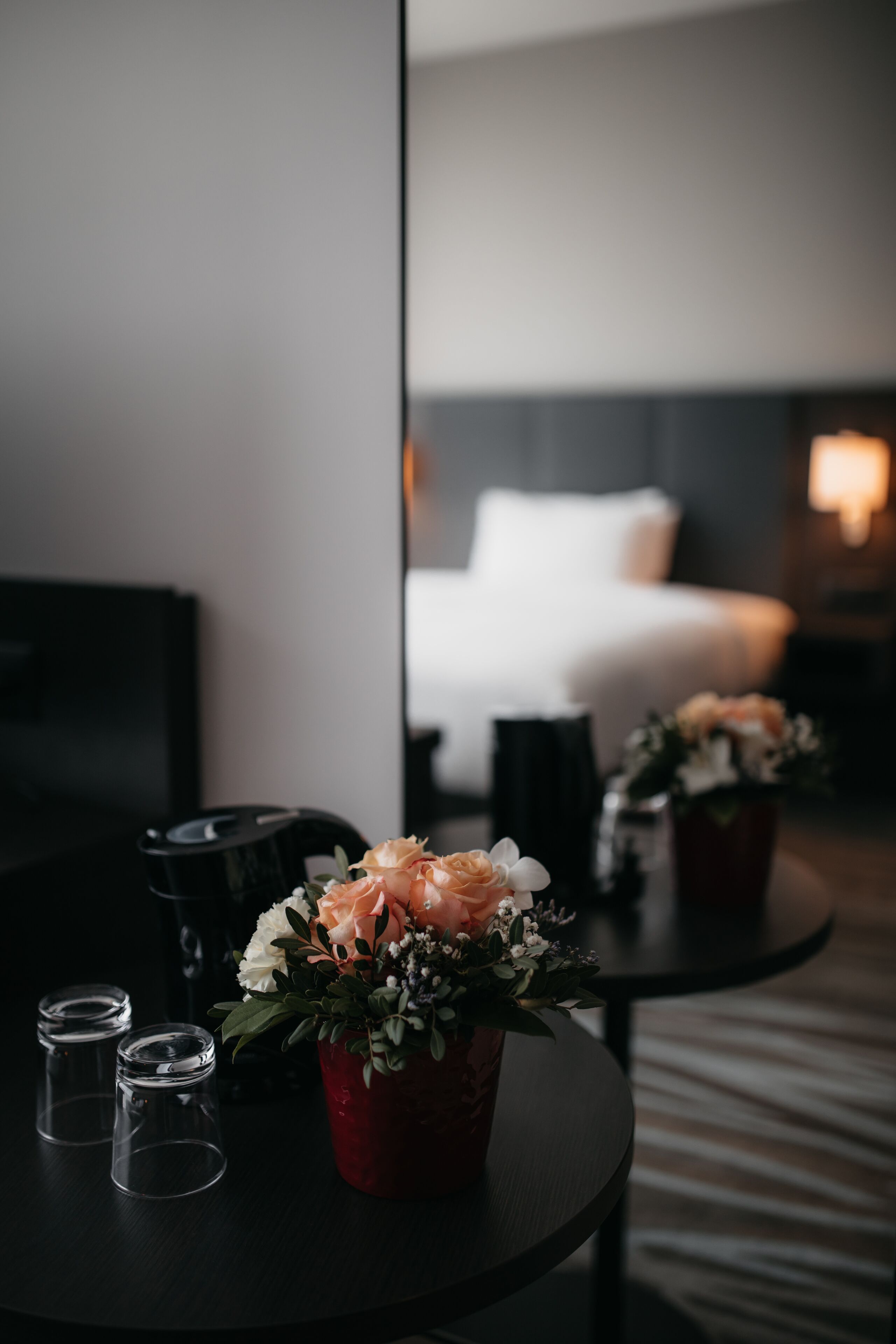 A hotel room with a bed, a bedside table with a vase of flowers, and a mirror reflecting the room.
