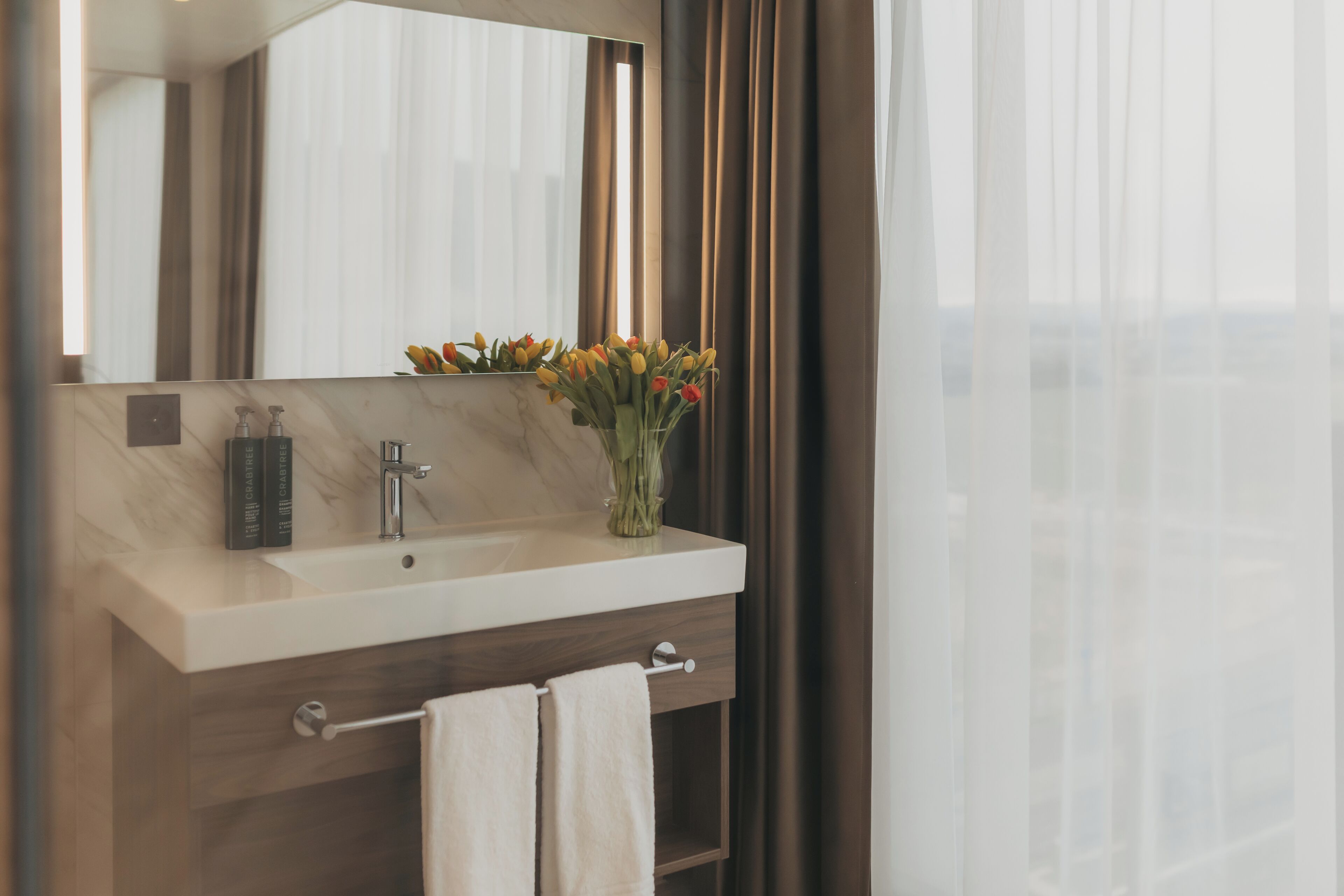 Un baño moderno con lavamanos de mármol, un espejo, un florero y un perchero para toallas.
