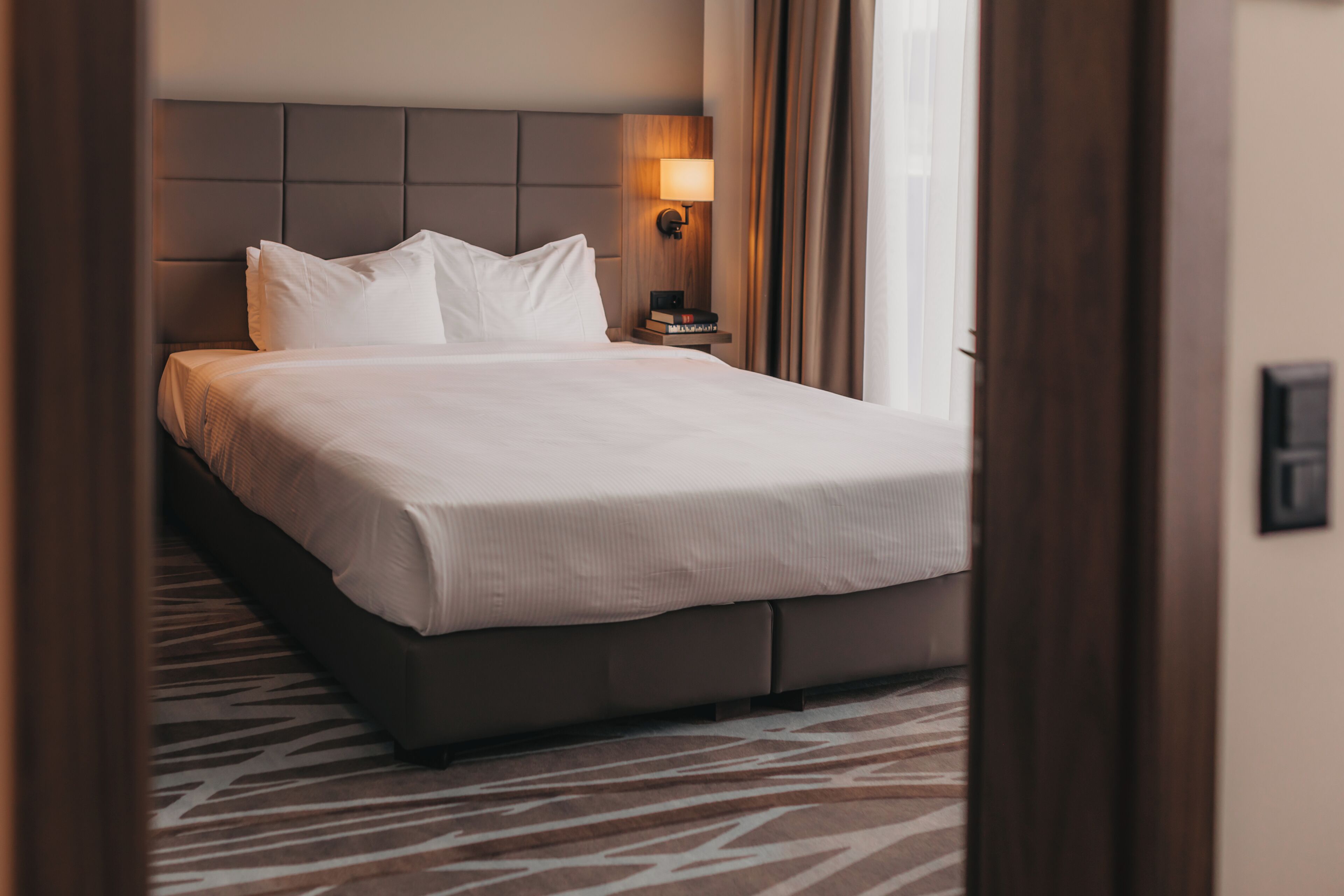 A neatly made bed with a headboard, a bedside table with a lamp, and a patterned carpet.