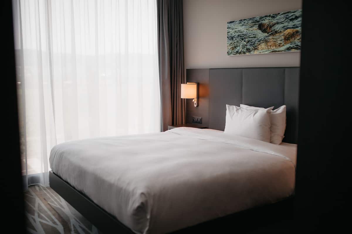 A hotel room with a large bed, a headboard, a nightstand with a lamp, a painting on the wall, and sheer curtains.
