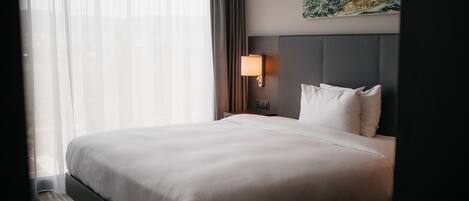 A hotel room with a large bed, a headboard, a nightstand with a lamp, a painting on the wall, and sheer curtains.