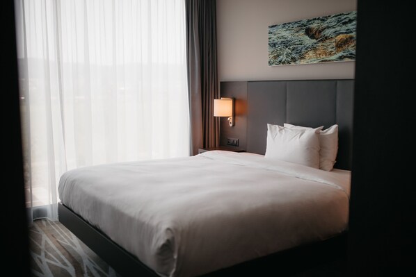 A hotel room with a large bed, a headboard, a nightstand with a lamp, a painting on the wall, and sheer curtains. - IIP Apartments - Self Check In (Spreitenbach)