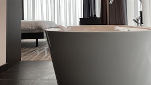 A modern bathroom with a freestanding bathtub, a vanity with a mirror, and a bed in the background. - IIP Apartments - Self Check In (Spreitenbach)
