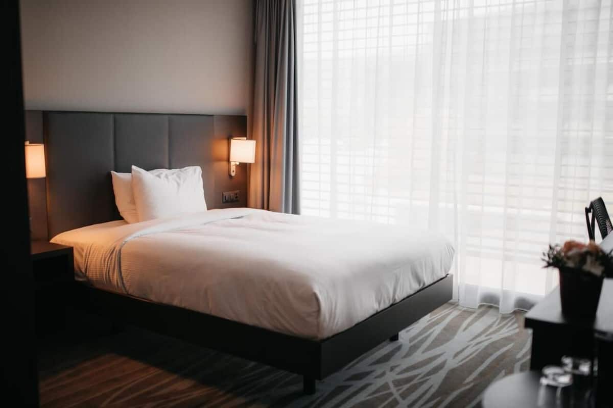 A modern hotel room with a large bed, a headboard with built-in lighting, a bedside table with a lamp, and a view through sheer curtains.