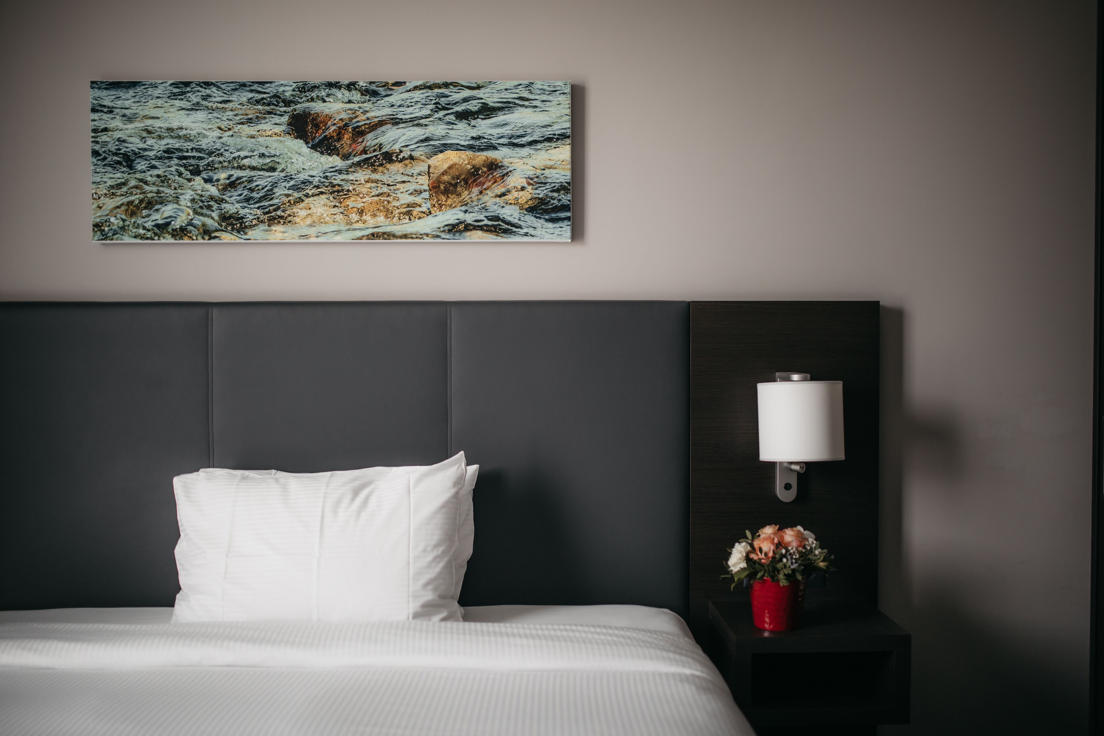 Quarto de hotel com cama, mesa de cabeceira com abajur, um vaso com flores e um quadro na parede.