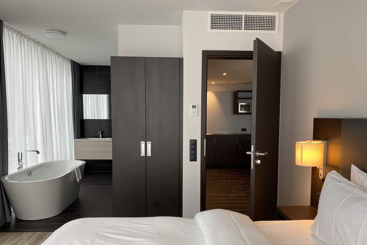 A modern hotel room with a large bed, a bathtub, a separate bathroom, and a kitchen area visible through an open door.