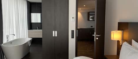 A modern hotel room with a large bed, a bathtub, a separate bathroom, and a kitchen area visible through an open door.