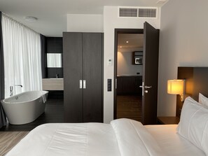 A modern hotel room with a large bed, a bathtub, a separate bathroom, and a kitchen area visible through an open door. - IIP Apartments - Self Check In (Spreitenbach)