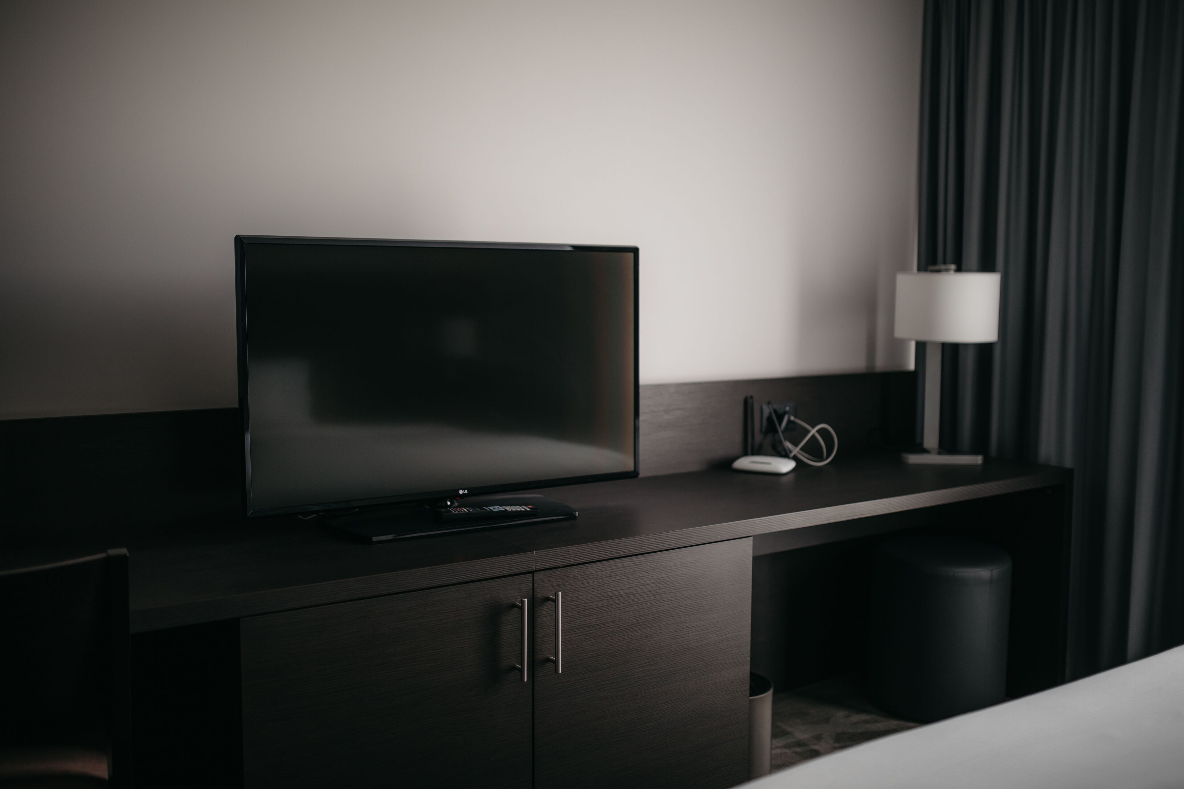 Une chambre d’hôtel moderne avec un téléviseur à écran plat posé sur un bureau en bois foncé, une lampe blanche et une poubelle.