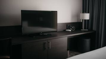 Une chambre d’hôtel moderne avec un téléviseur à écran plat posé sur un bureau en bois foncé, une lampe blanche et une poubelle.