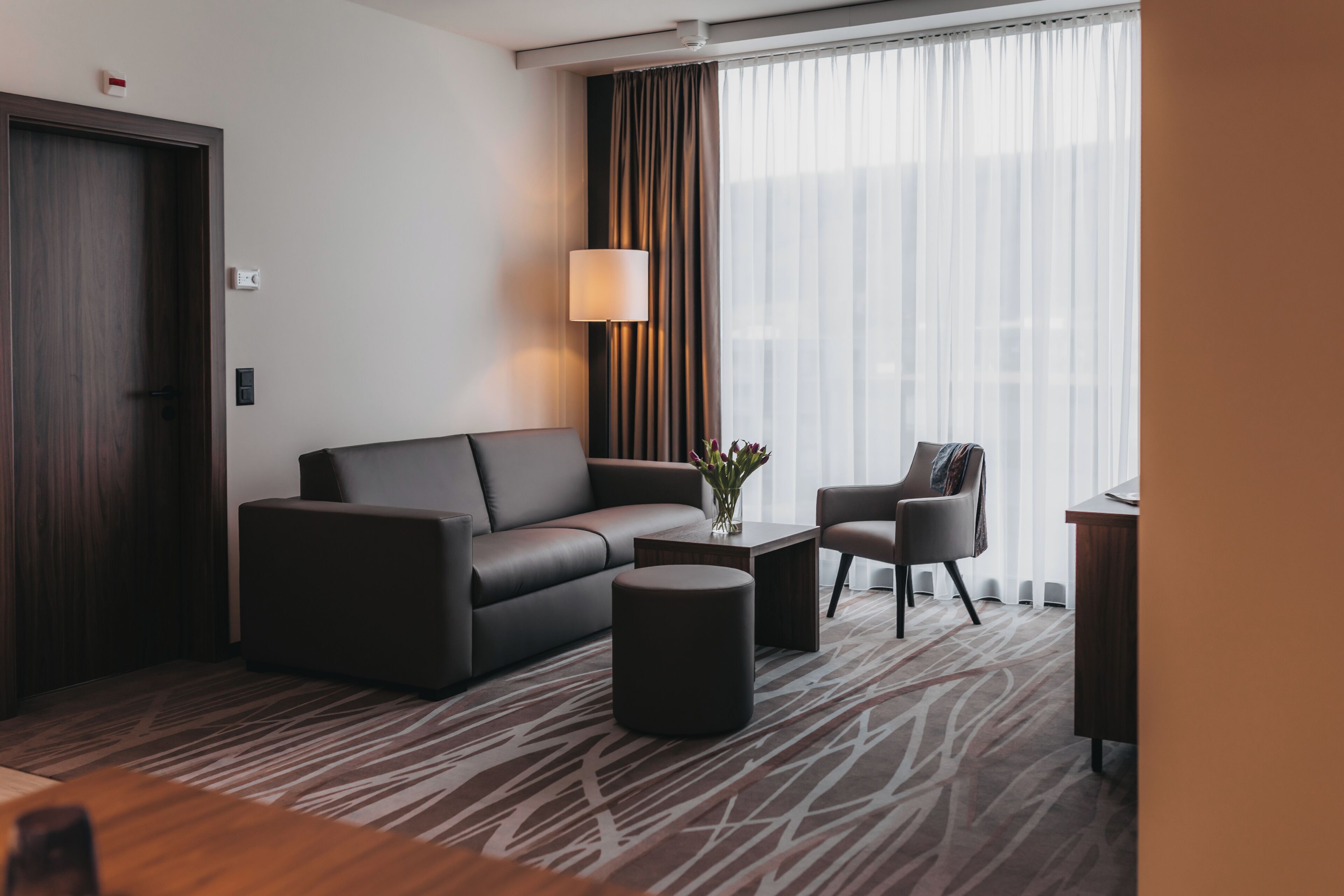 Et moderne hotelværelse med en mørkebrun sofa, en lille rund fodskammel, et sofabord med en vase og en stol.