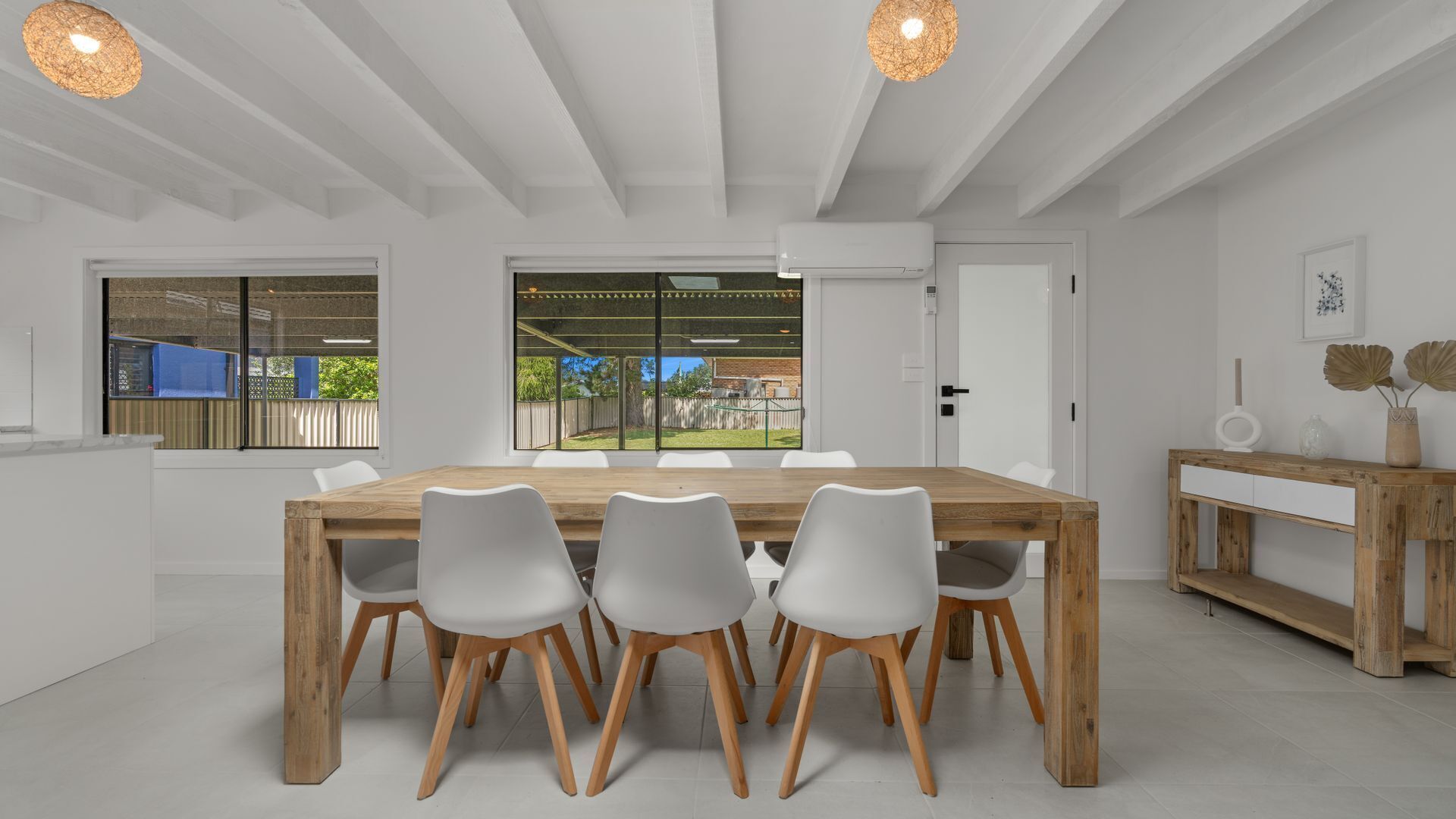 A modern dining area with a wooden table and white chairs, large windows offering a view of the outdoors, and a minimalist decor with a wooden console table and a vase.