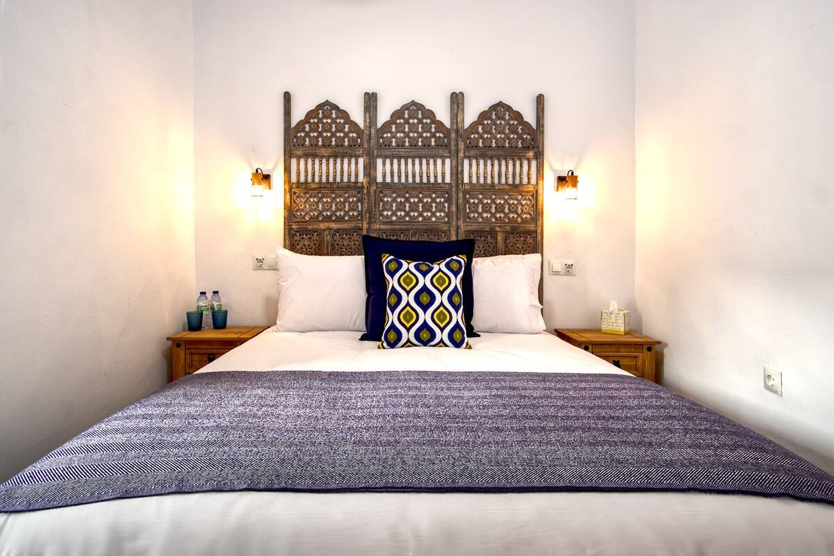 Ein ordentlich gemachtes Bett mit einem gemusterten Überwurf, zwei Kissen und einem dekorativen Kopfteil mit Wandleuchten.