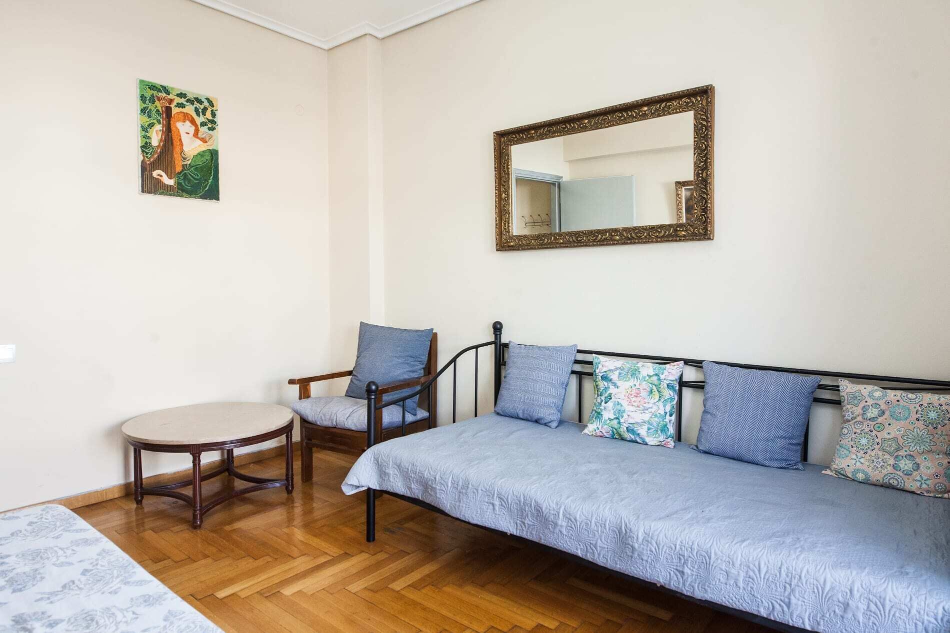 A room with a wooden floor, a round table, a sofa with patterned cushions, a chair, a mirror with an ornate frame, and a wall-mounted picture.