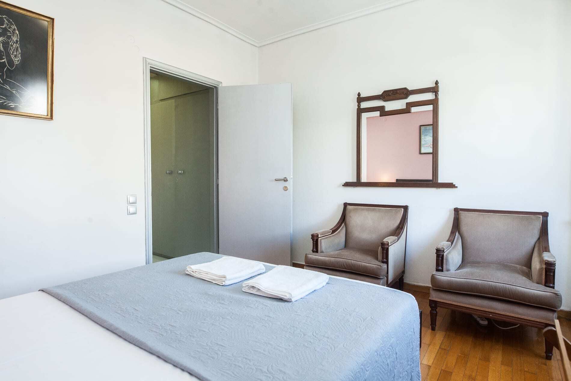 A bedroom with a bed, two armchairs, a door, and a mirror.