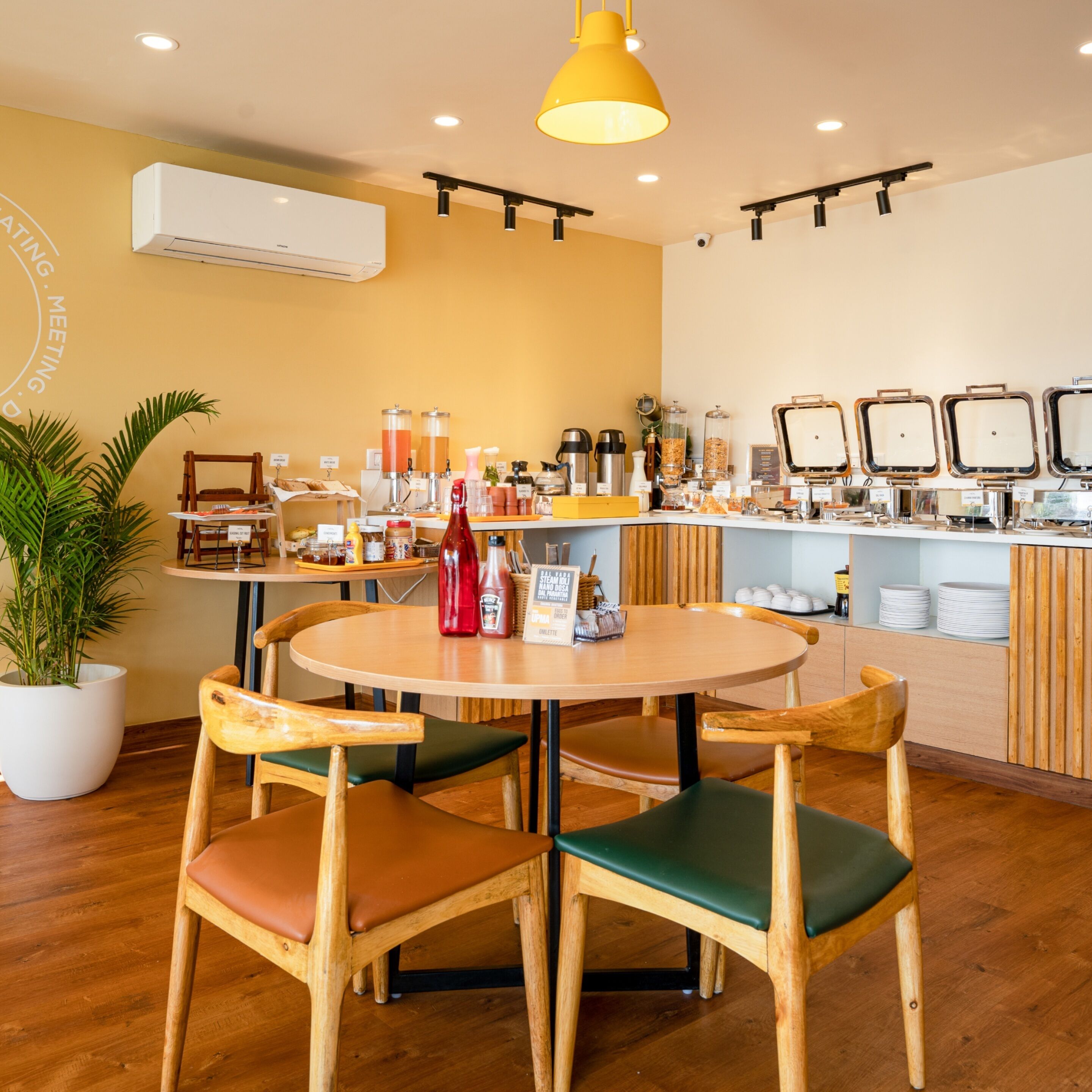 Um restaurante com uma mesa e cadeiras de madeira redondas, uma luminária pendente amarela e um balcão com diversos alimentos e acompanhamentos.