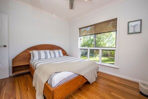 A bedroom with a wooden bed, a window with a view of greenery, and a ceiling fan. - The Wave (Horseshoe Bay)