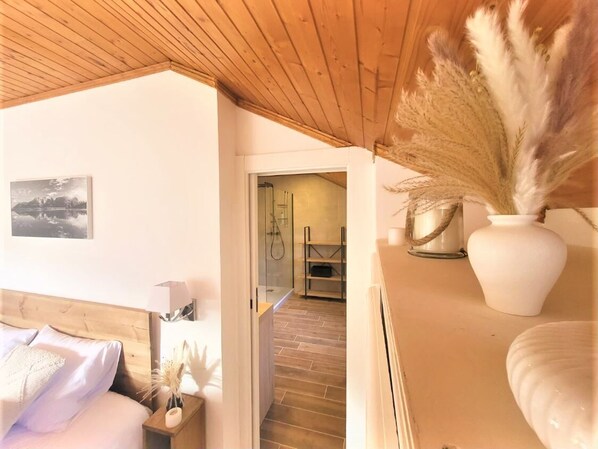 Un dormitorio con techo de madera, una cama con almohadas, una mesita de noche con una lámpara, un jarrón con plumas de pampa y un cuadro enmarcado en la pared.