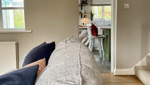 A living room with a grey sofa, a window with blinds, a wall-mounted light, and a view into the kitchen area.