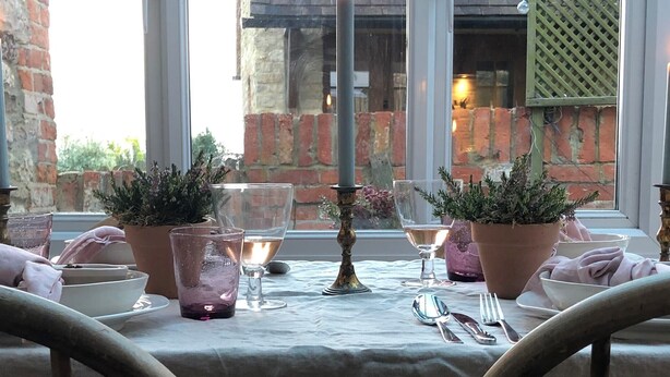 A table set with candles, glassware, and potted plants, with a view of a brick wall and window in the background.
