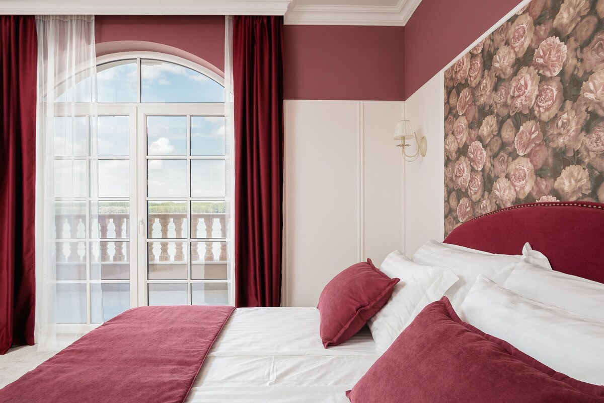 Une chambre avec un grand lit, des rideaux rouges, un balcon avec vue et une fresque murale à motifs floraux.