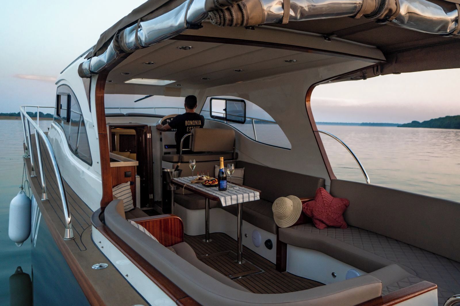 A boat interior with a dining area, a person standing by the window, and a view of the water.