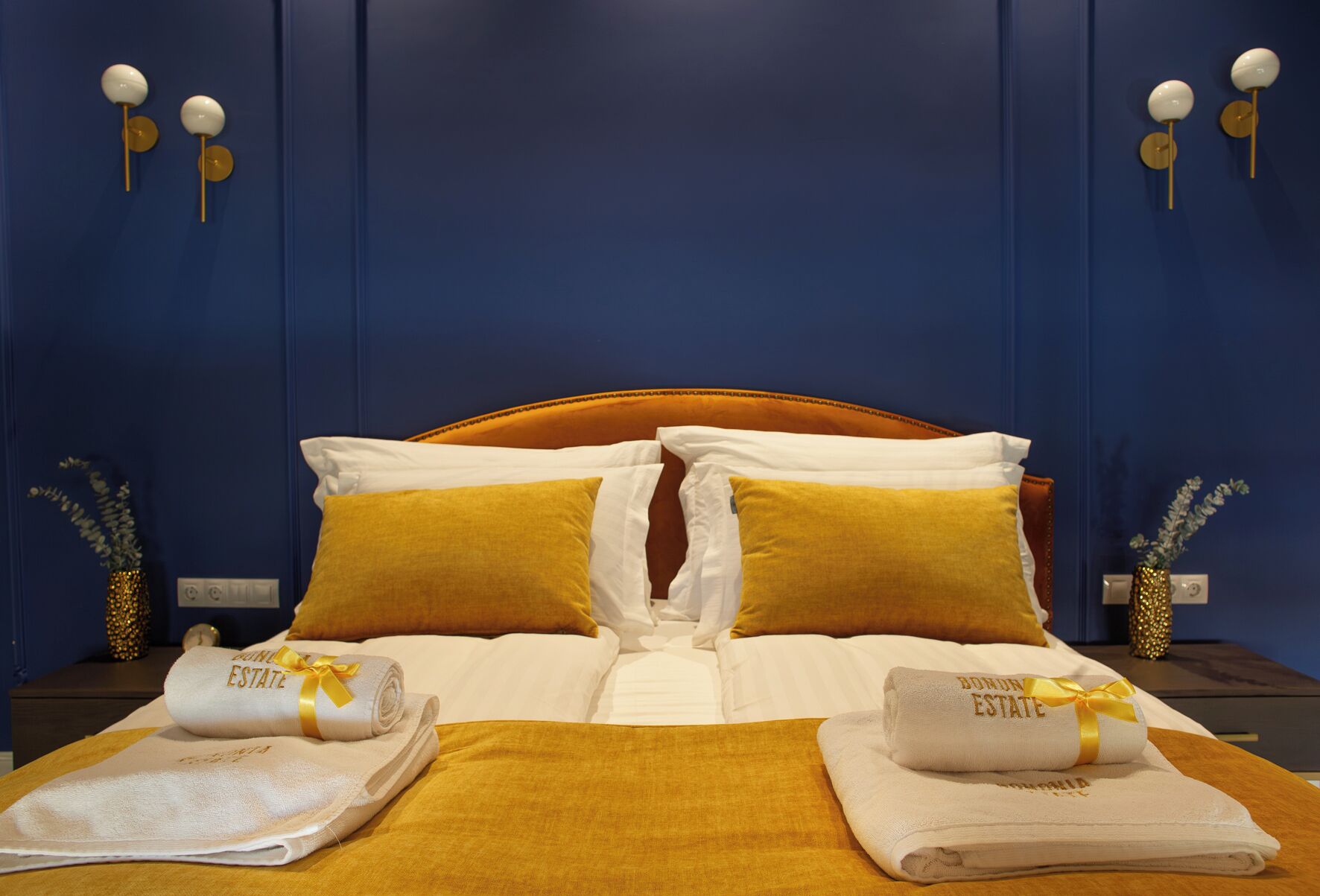 Una cama bien tendida con almohadas blancas y mostaza, un detalle dorado y una toalla doblada con la inscripción "Bonn ESTATE".