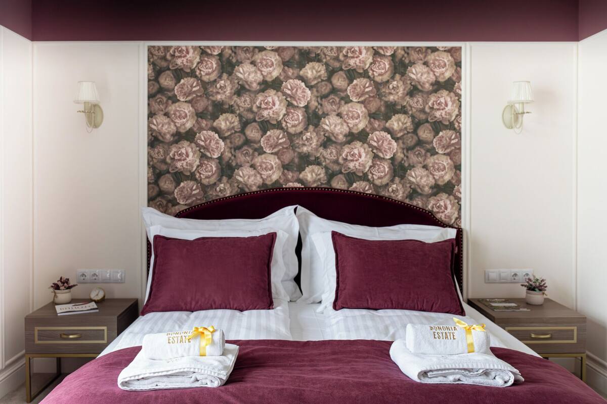 Un lit bien fait, avec une tête de lit recouverte d’un papier peint à motifs floraux, deux oreillers blancs, deux oreillers bordeaux et deux ensembles de serviettes ornés d’un ruban jaune.