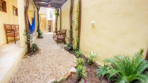 Un sendero estrecho con superficie de grava, bordeado por plantas y sillas de madera, que conduce a una zona cubierta con una pérgola.