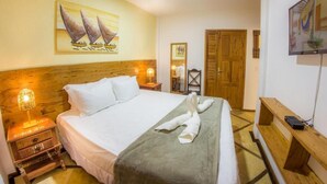 Habitación de hotel con cama, cabecera de madera, un cuadro de veleros, un banco de madera, televisor y puerta de madera.