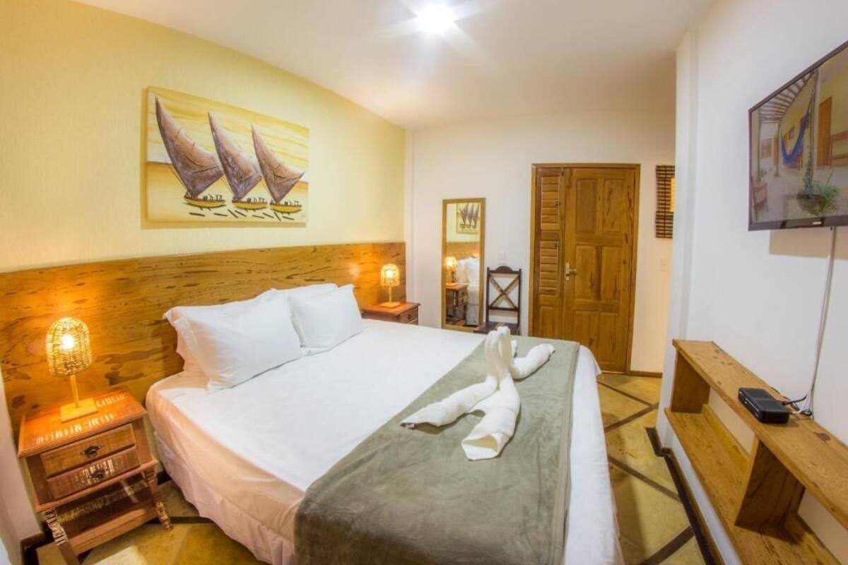 A hotel room with a bed, wooden headboard, a painting of sailboats, a wooden bench, a TV, and a wooden door.