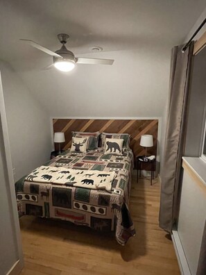 Ein Schlafzimmer mit einem Bett, Deckenventilator, Nachttisch, Lampe und einem Fenster mit Vorhängen.
