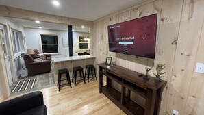 A modern living room with a large flat-screen TV mounted on the wall, a wooden dining area, and a cozy seating area with a sofa and bar stools.