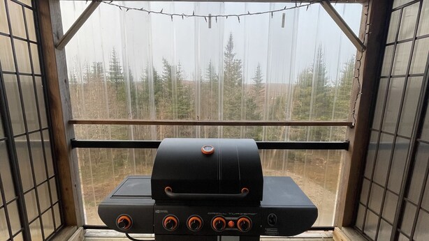 A barbecue grill with four burners, a control panel, and a digital display, set on a wooden deck with a covered roof and a view of trees.
