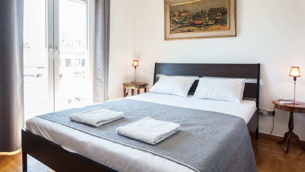 A hotel room with a large bed, two bedside tables, a painting on the wall, and a view through a window. - Gorgeous 2 bedrooms apt at Exarcheia (Athens)