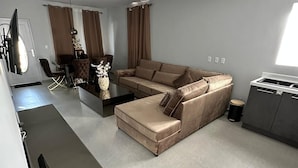 A modern living room with a beige sectional sofa, a dining area with a table and chairs, and a kitchen area with a grey cabinet. - Beautiful detached home with pool and gym! (Willemstad)