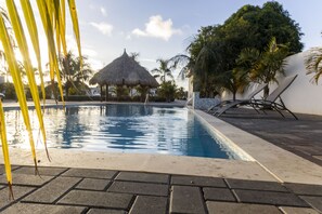 A swimming pool with a thatched roof structure in the background, surrounded by palm trees and a paved walkway.