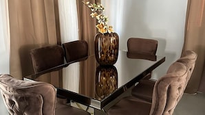 A modern dining area with a glass-top table, four cushioned chairs with gold legs, and a vase with flowers. - Beautiful detached home with pool and gym! (Willemstad)