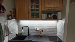 A compact kitchen with wooden cabinets, a black sink, a built-in washing machine, and a window with blinds. - Studio-cabin apartment (Combloux)