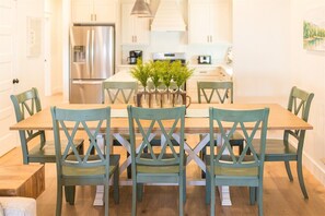 A dining area with a wooden table and four chairs, a kitchen in the background, and a vase with greenery on the table. - Brown Eyed Girl at River Rocks Plantation (Guntersville)
