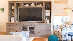 Ein Wohnzimmer mit großem Flachbildfernseher, einer Holz-Unterhaltungseinrichtung, einem gemusterten Sessel und einem runden, grünen Hocker.