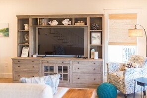 A living room with a large flat-screen TV, wooden entertainment center, patterned armchair, and a green round ottoman. - Brown Eyed Girl at River Rocks Plantation (Guntersville)