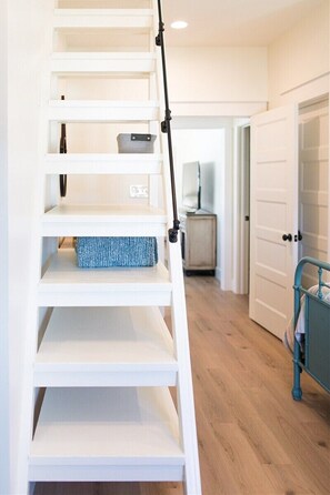 Una escalera con escalones blancos y barandilla negra que conduce a una habitación con una cama y una mesita de noche.