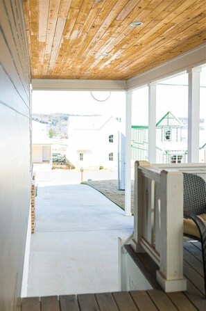 A covered porch with a wooden ceiling, white walls, and a view of a white building in the background. - Brown Eyed Girl at River Rocks Plantation (Guntersville)
