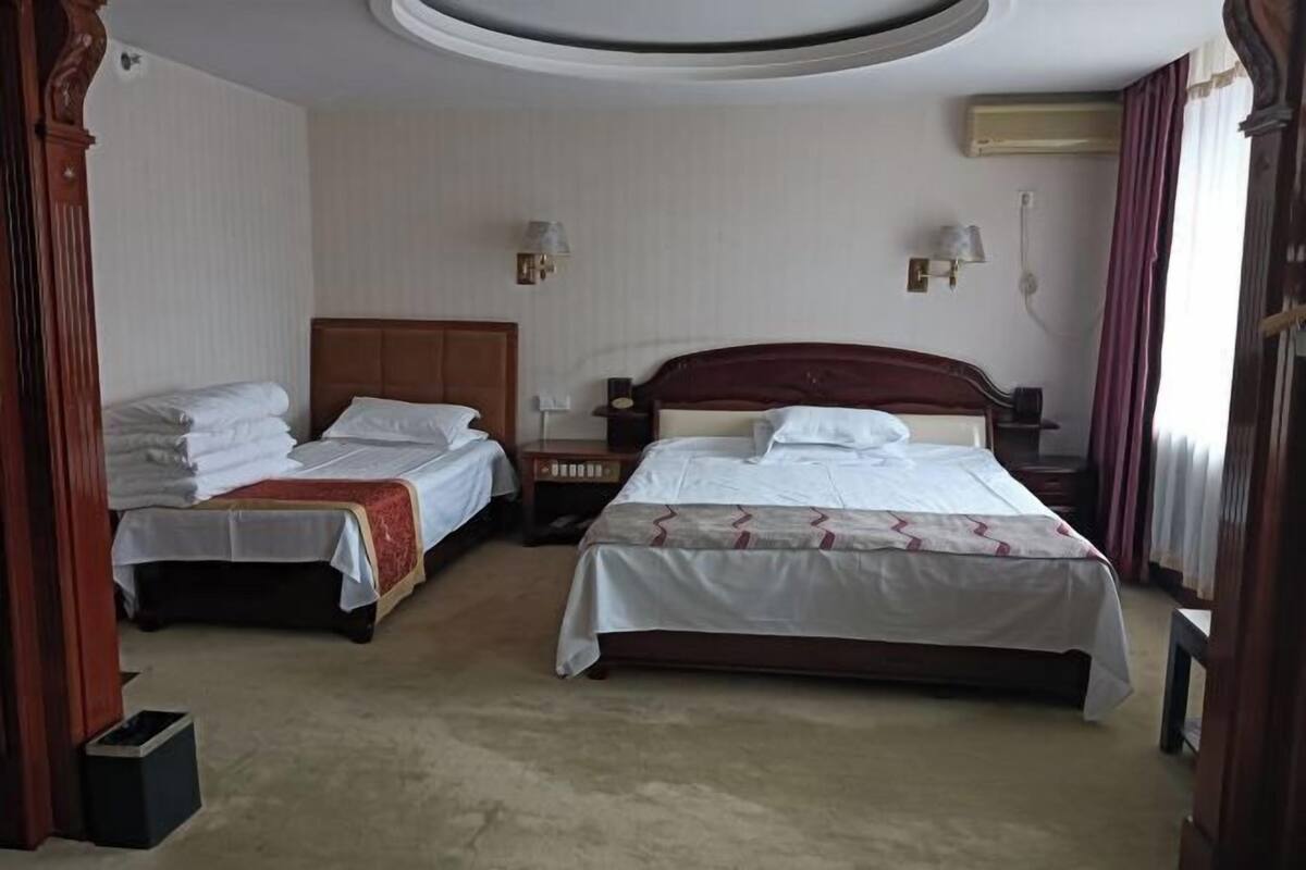 Habitación de hotel con dos camas, cabecera de madera, mesita de noche pequeña, lámpara de pared y lámpara en el techo.