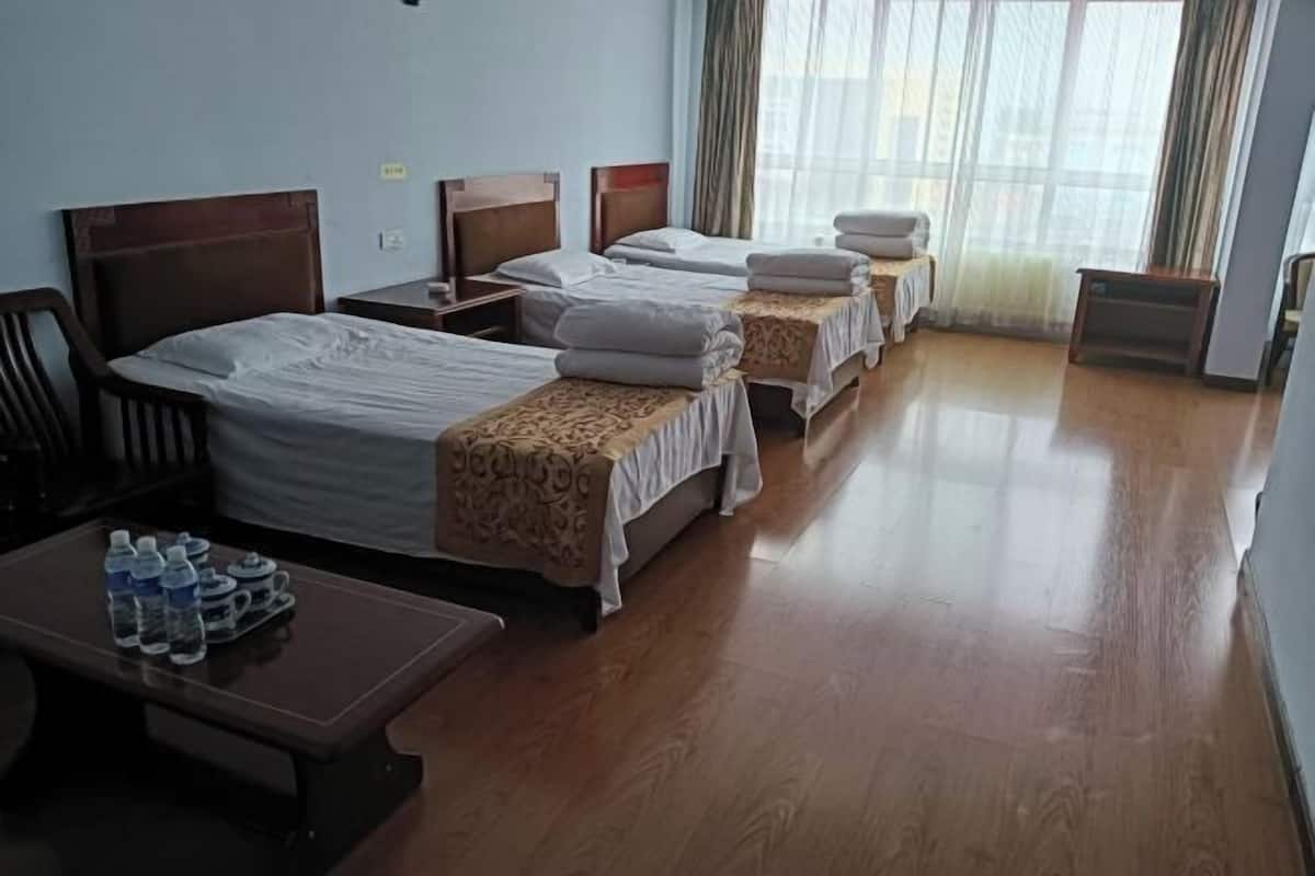 Habitación de hotel con tres camas, piso de madera, un escritorio y botellas de agua, y una ventana con cortinas.