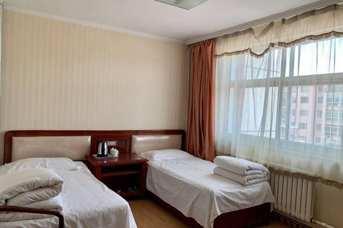 Habitación de hotel con dos camas individuales, una mesita de noche de madera con hervidor y una ventana con cortinas translúcidas.