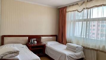 A hotel room with two single beds, a wooden nightstand with a kettle, and a window with sheer curtains.
