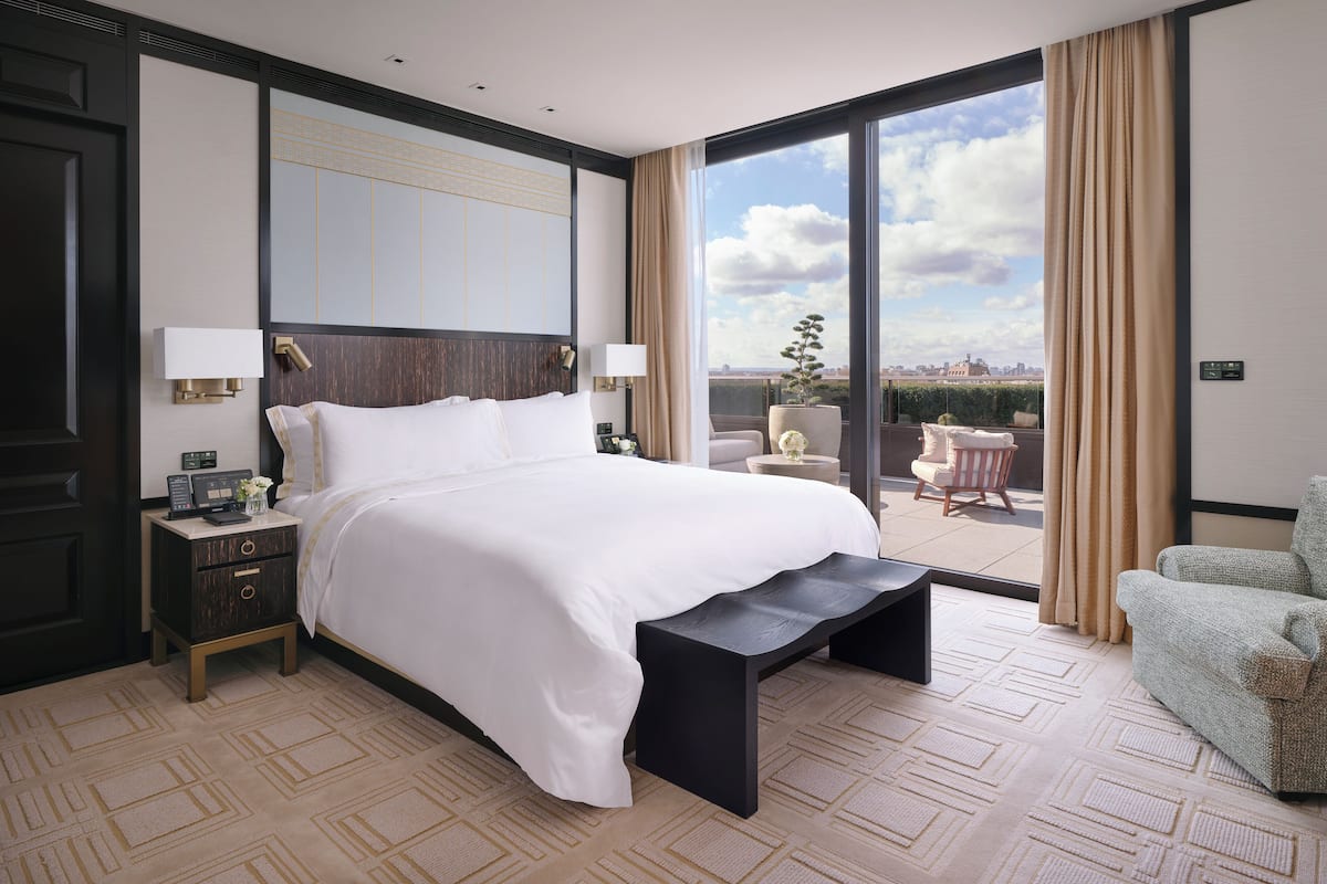 A modern hotel room with a large bed, a nightstand, a bedside table, a chair, and a view of the city through large windows.