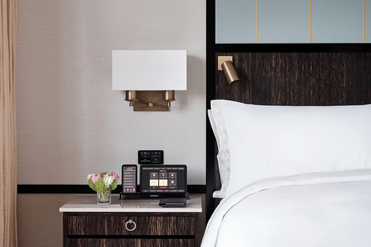 A hotel room with a bed, a bedside table with a lamp, a thermostat, and a vase with flowers.